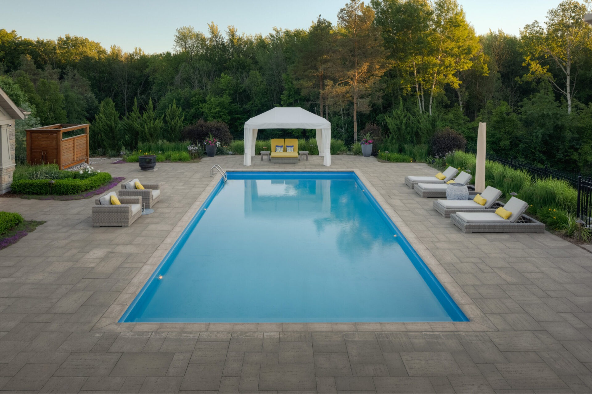 An inviting outdoor swimming pool with loungers, a white canopy, and a surrounding patio set within a lush landscape during the early evening.