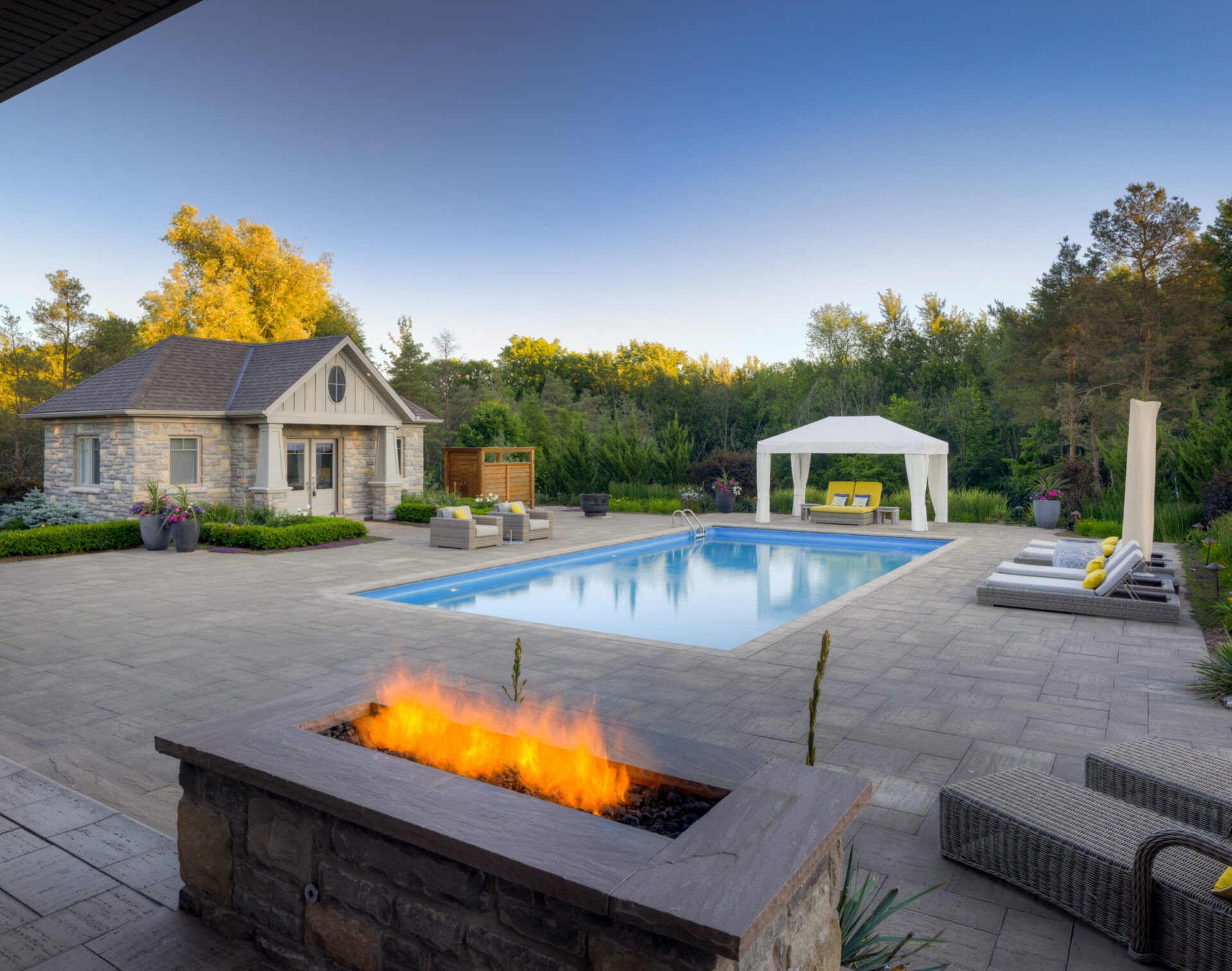A luxurious backyard with an in-ground pool, elegant stone house, outdoor seating, a fire pit, and lush greenery under a clear, twilight sky.