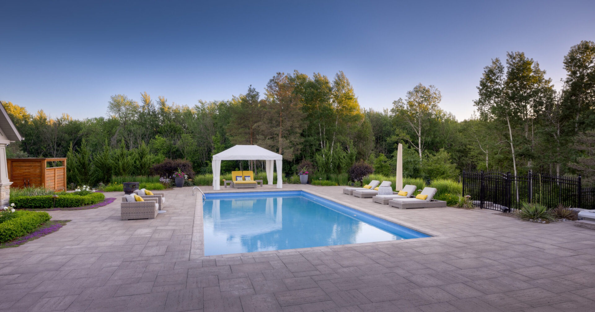 A serene backyard with a rectangular swimming pool, patio furniture, a white gazebo, surrounded by a lush forest and a clear sky above.