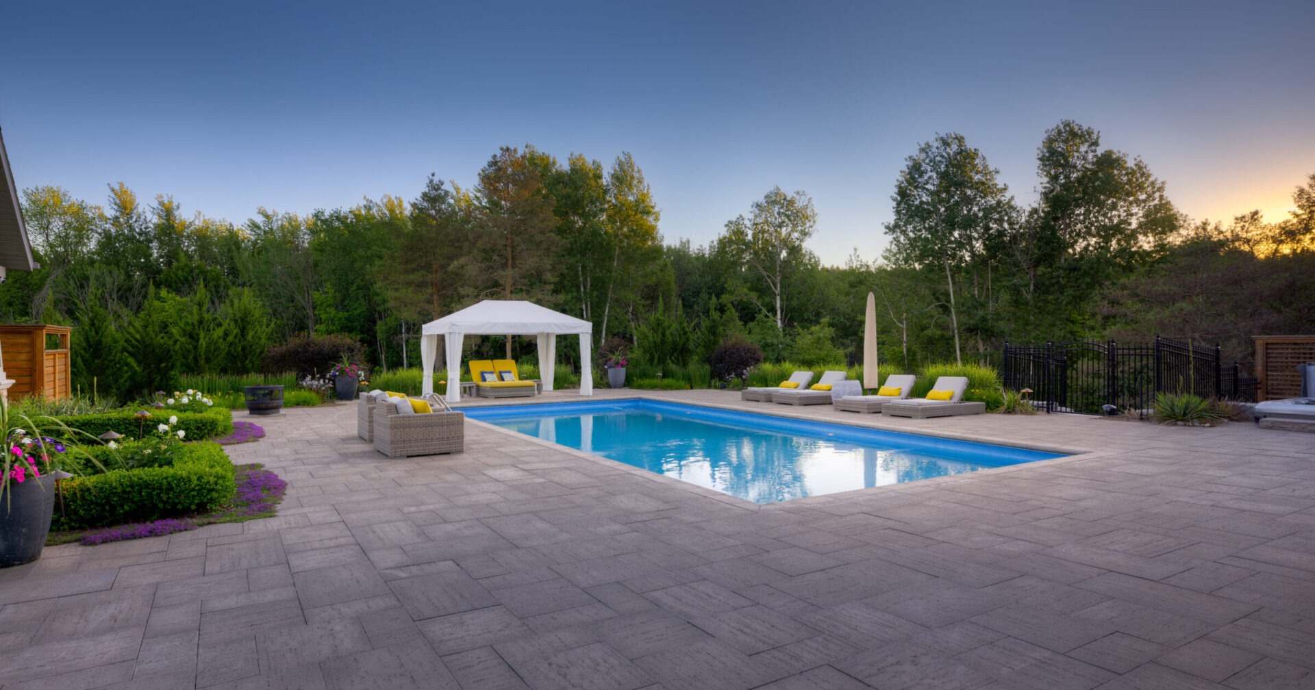 Luxurious backyard with a rectangular pool, patio, loungers, gazebo, and garden at twilight, surrounded by lush trees and a clear sky.