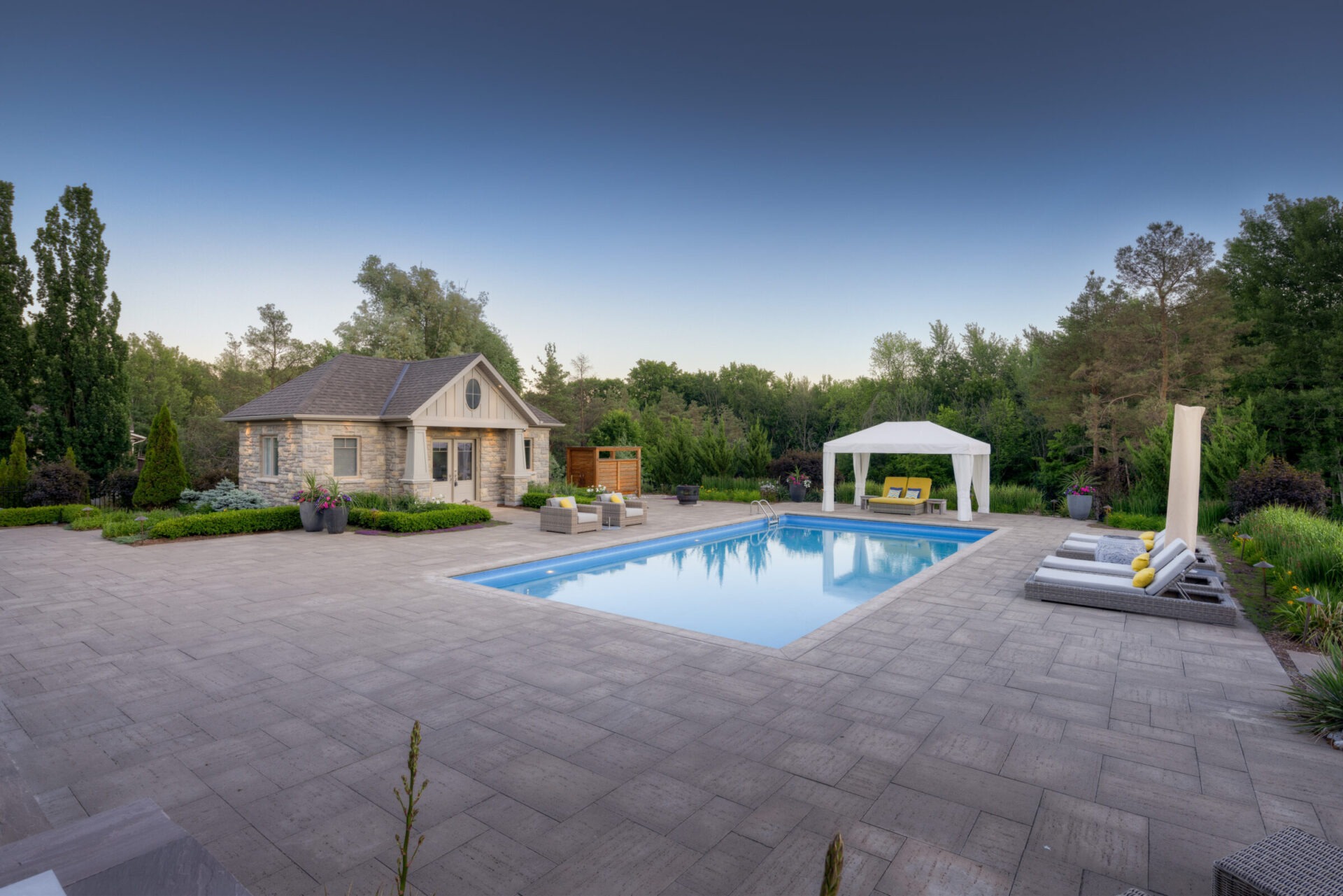 A tranquil backyard setting with a swimming pool, stone patio, cozy pool house, verdant garden, gazebo, and clear blue sky at dusk.