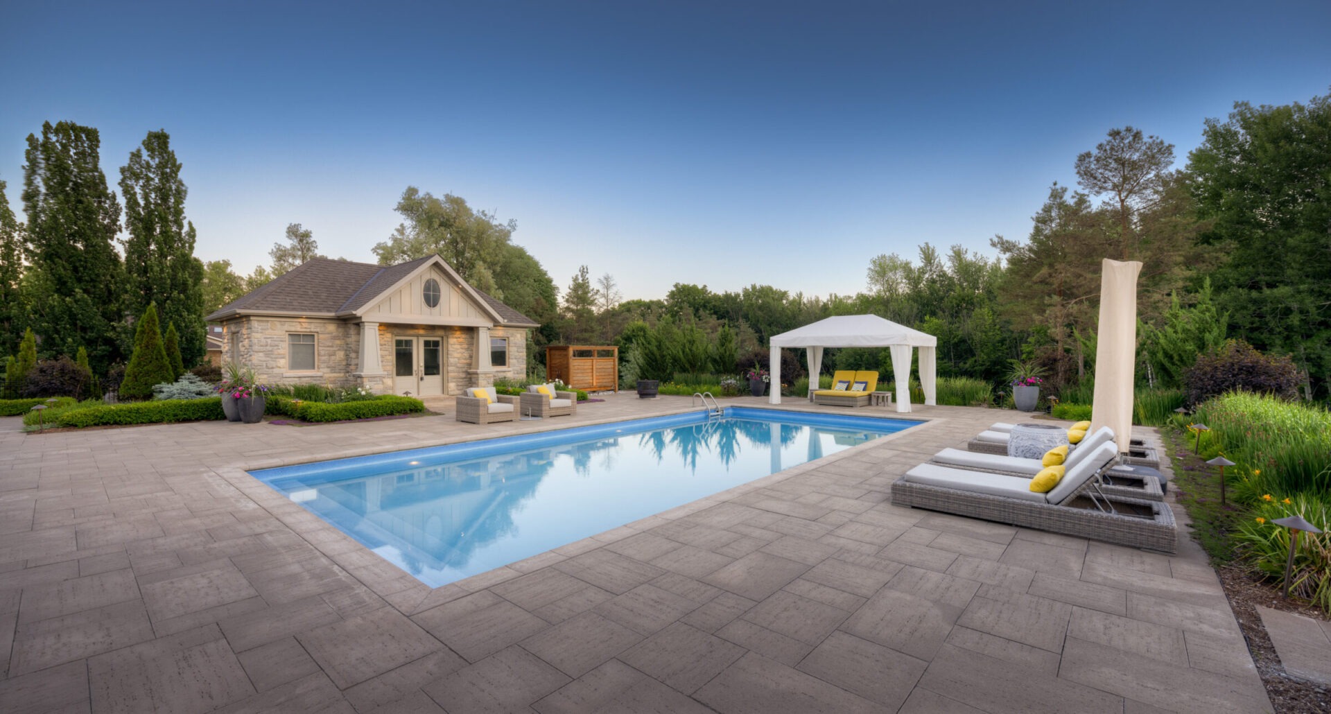 A tranquil backyard setting featuring a clear swimming pool, elegant loungers, a gazebo, lush landscaping, and a stone-clad house under a blue sky.