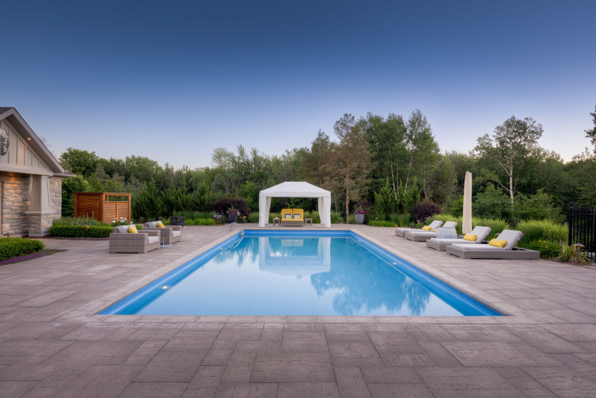 An outdoor swimming pool with a gazebo, lounge chairs, surrounding greenery, and a stone patio during twilight in a tranquil backyard setting.