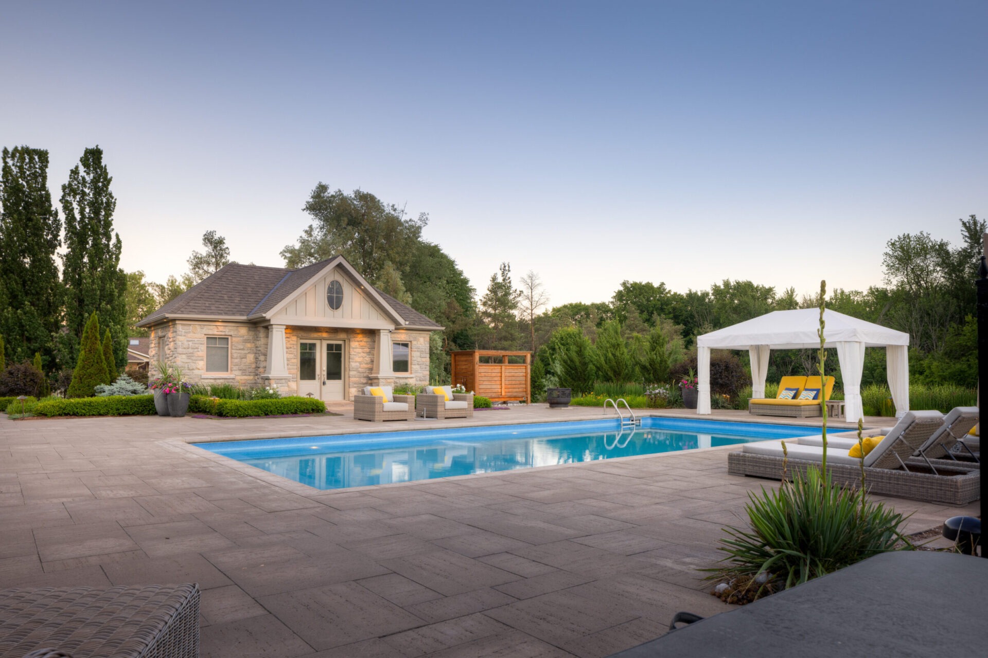 A serene backyard with a swimming pool, patio, poolside cabana, comfortable outdoor furniture, and a stone house surrounded by lush greenery at dusk.
