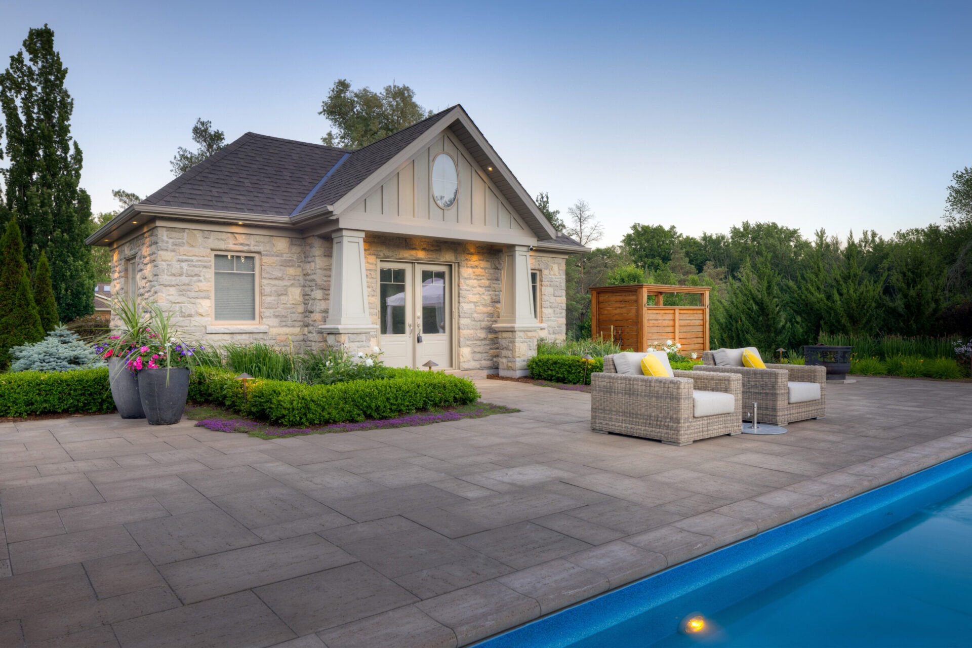 A cozy suburban house with stone veneer under a clear sky. A patio features outdoor furniture near a pool, surrounded by lush landscaping.