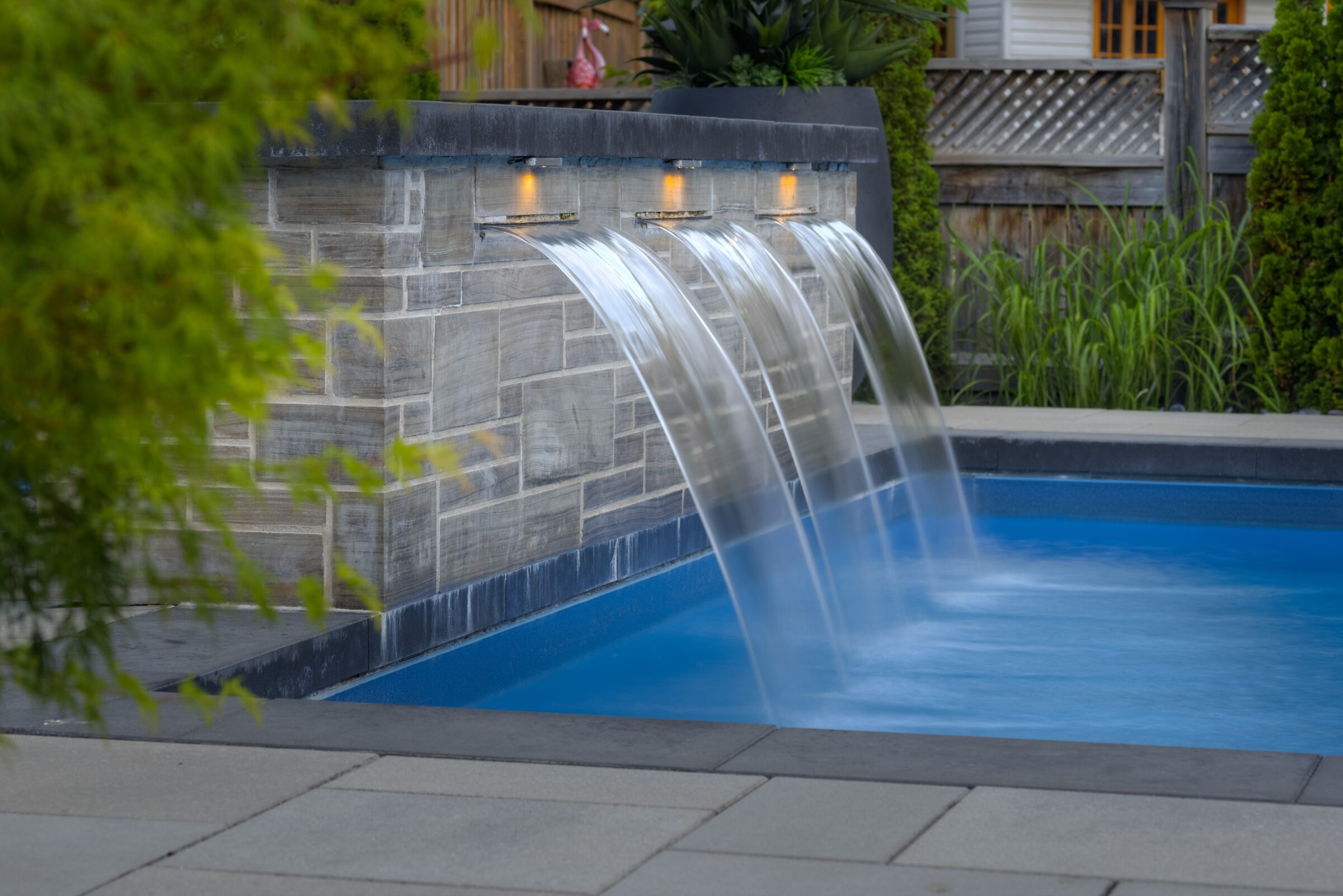 A serene backyard featuring a pool with a stone waterfall feature. Three arcs of water flow smoothly into the blue water, illuminated by overhead lights.