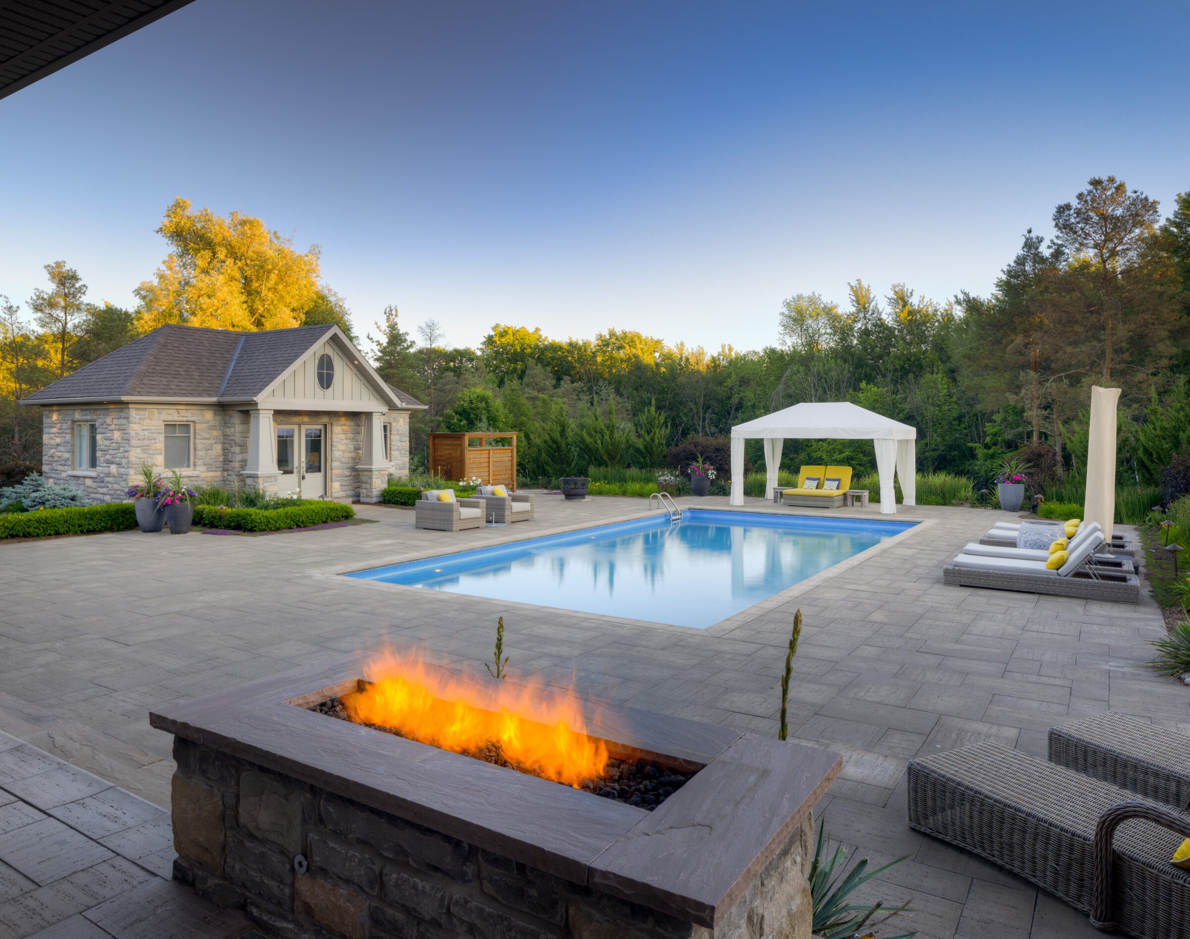 A luxurious backyard with a large swimming pool, gazebo, outdoor furniture, and a fire feature, flanked by a lush forest and a stone-clad house.