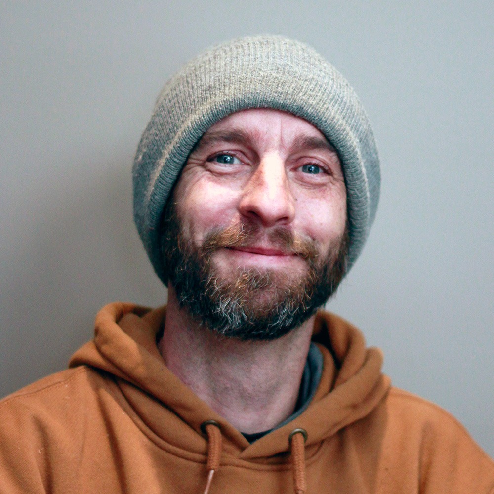 This image shows a smiling person with a beard, wearing a brown hoodie and a grey knitted hat, standing against a plain background.
