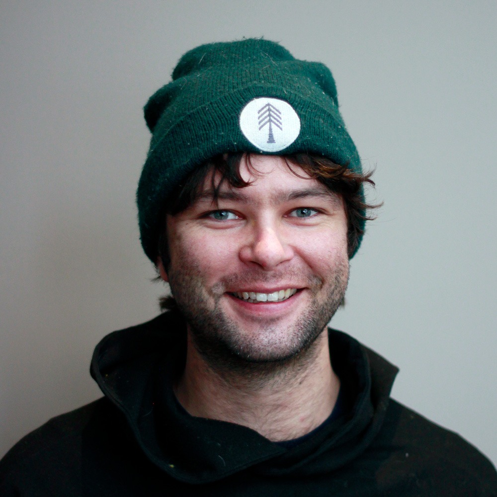 A smiling person wearing a green knitted hat with a tree emblem is in front of a neutral background. They have tousled hair and stubble.
