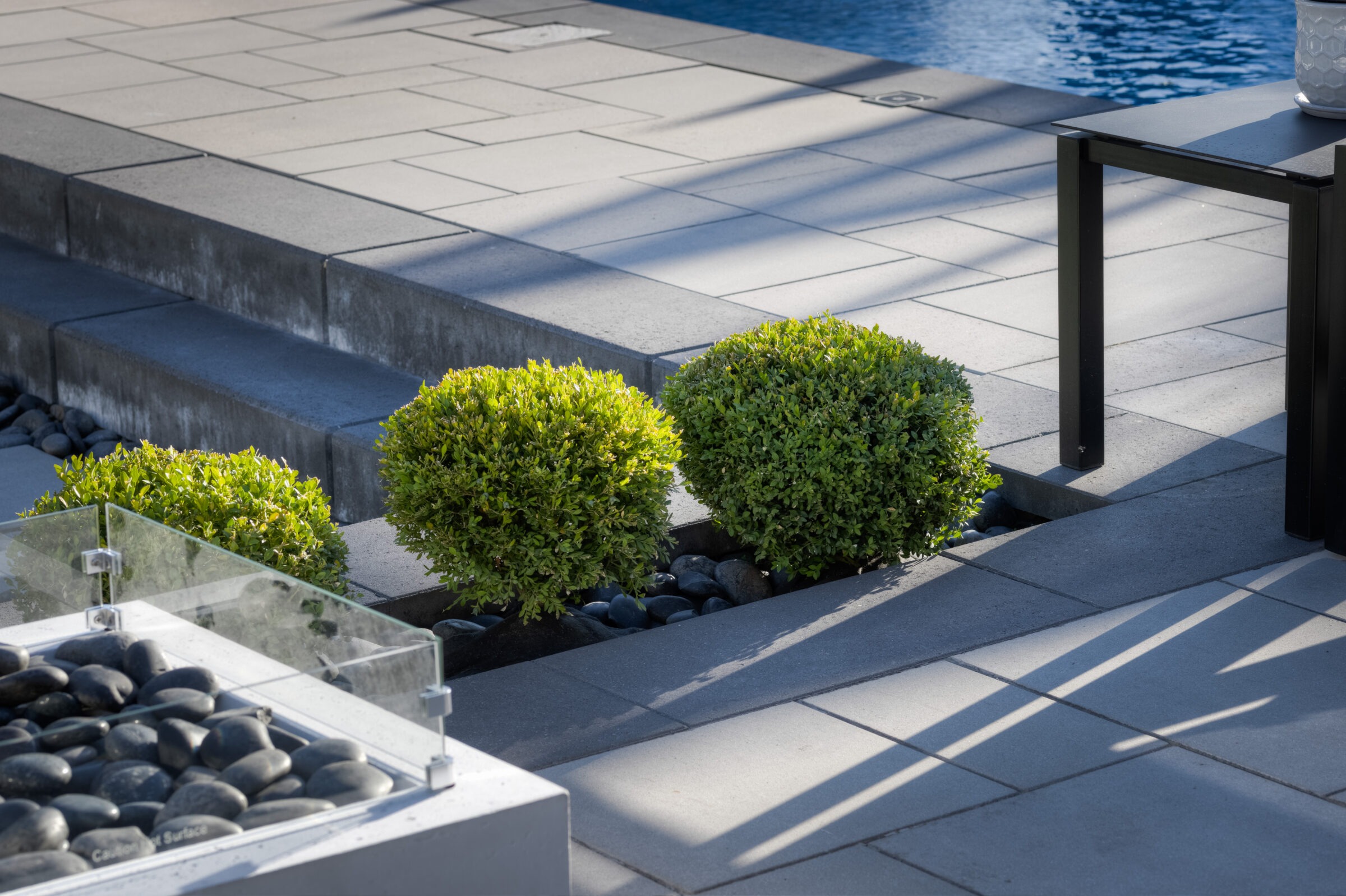 The image shows a modern outdoor space with neatly trimmed bushes, smooth paving stones, a glass barrier, and shadows indicating sunny weather.