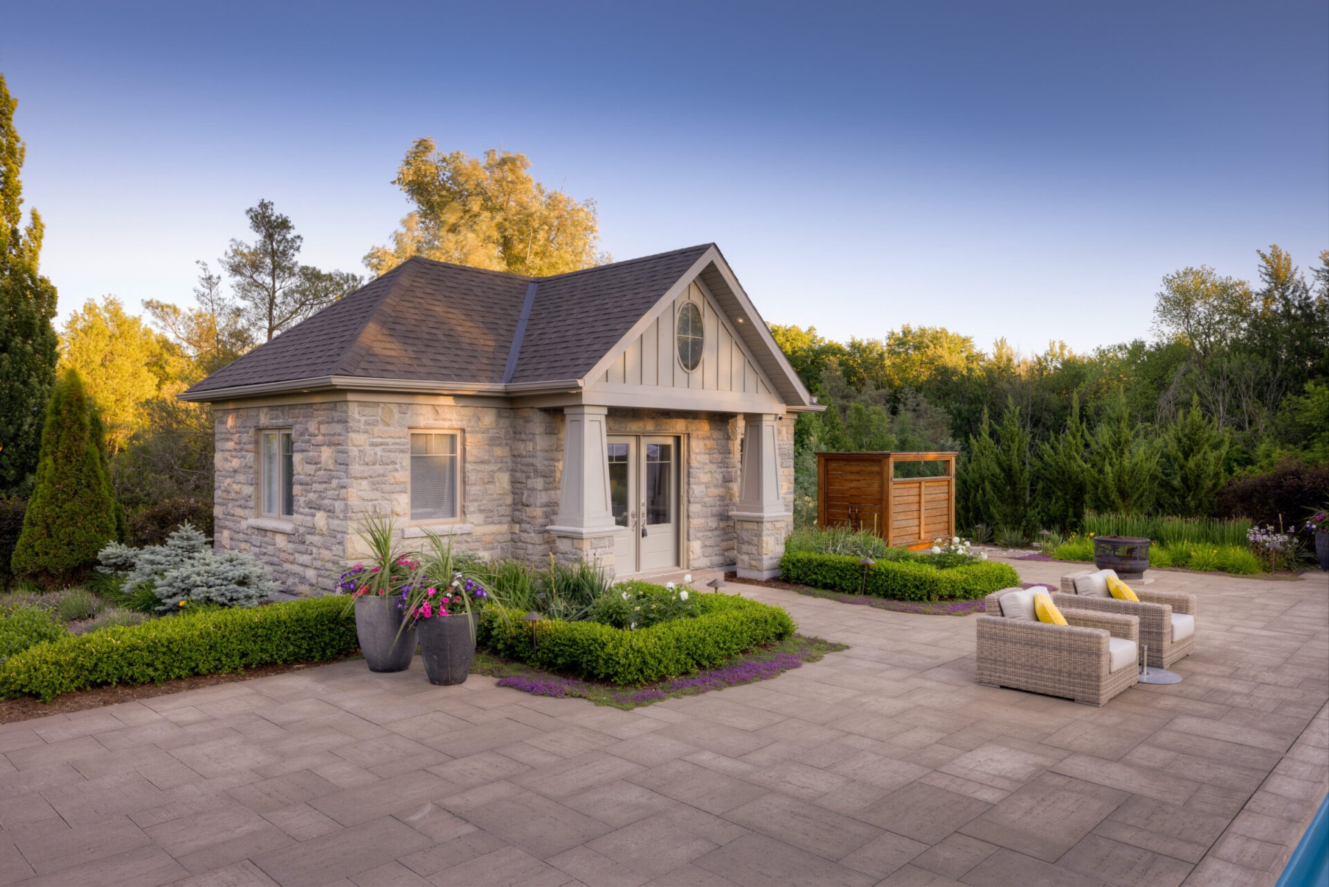 A quaint stone house with a gabled roof, flanked by lush gardens and outdoor furniture on a paver patio, set against a backdrop of trees at dusk.