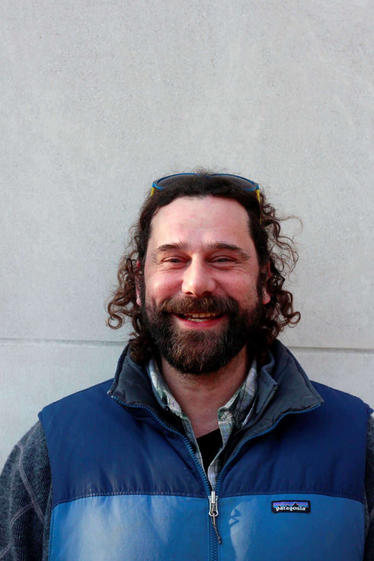 A smiling person with curly hair and a beard stands before a gray wall, wearing sunglasses atop their head and a two-toned blue vest.