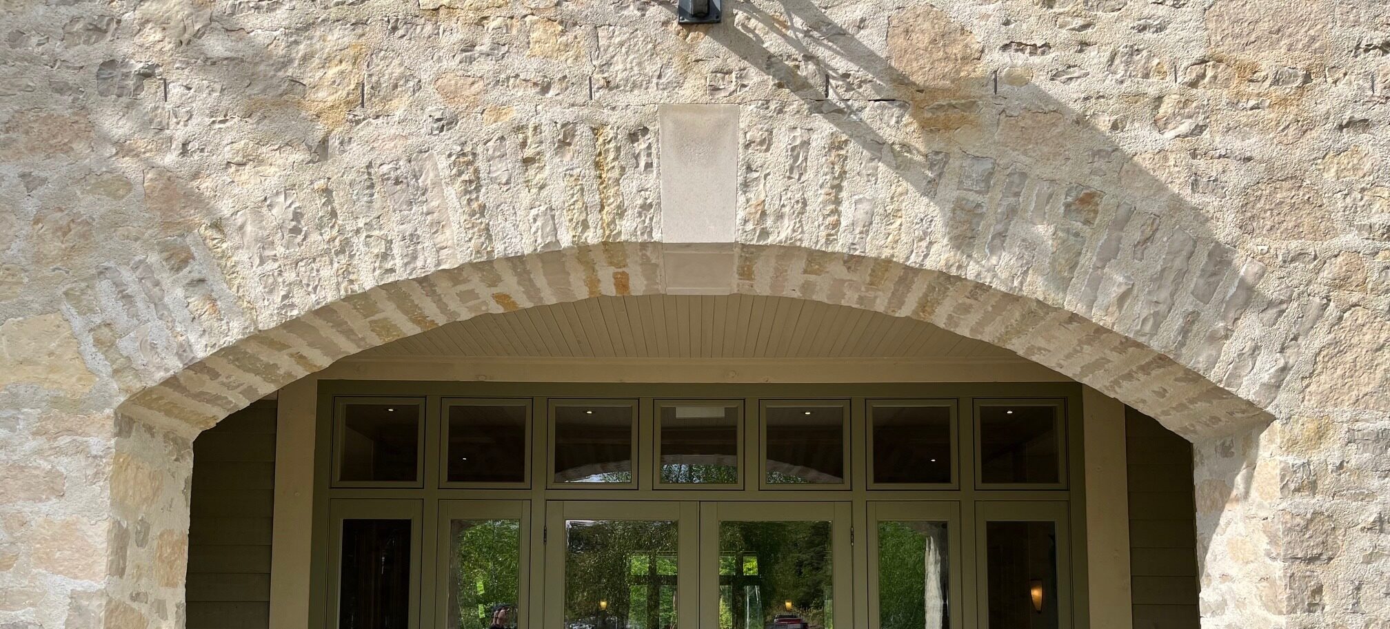 This image shows a stone archway above a set of modern glass doors, with sunlight casting shadows, creating a blend of historic and contemporary architectural styles.