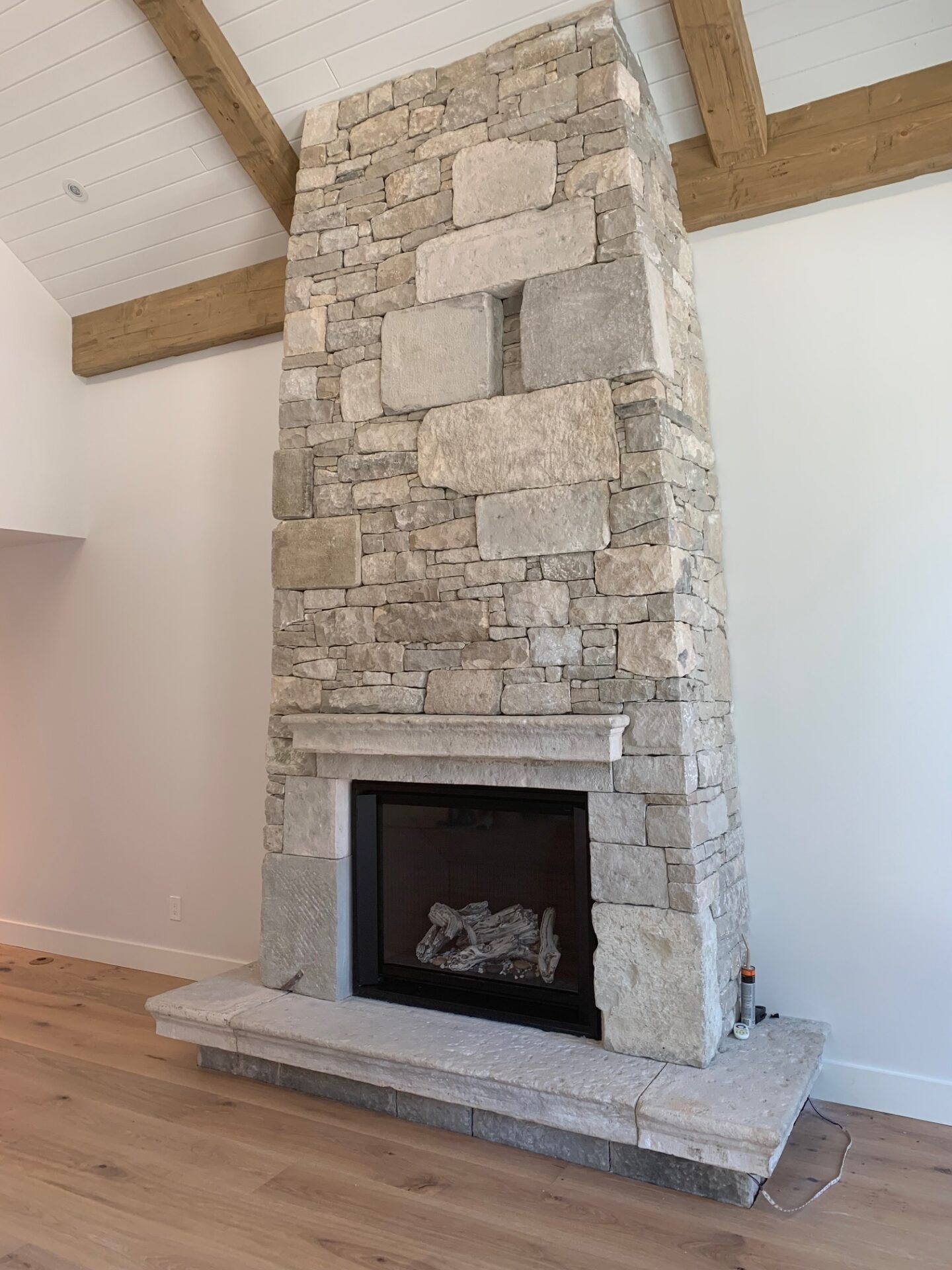 A spacious room with a high ceiling and wooden beams, featuring a large stone fireplace with a closed gas insert and a hardwood floor.
